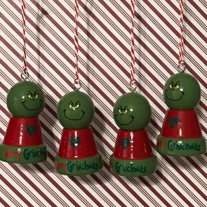 May include: Four green Grinch ornaments with red and green accents. Each ornament has a green Grinch face with a red heart and the text "Merry Grinchmas".