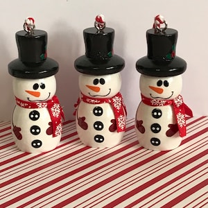 May include: Three small wooden snowman ornaments with black top hats, red and white scarves, and black buttons. The snowmen are on a red and white striped background.