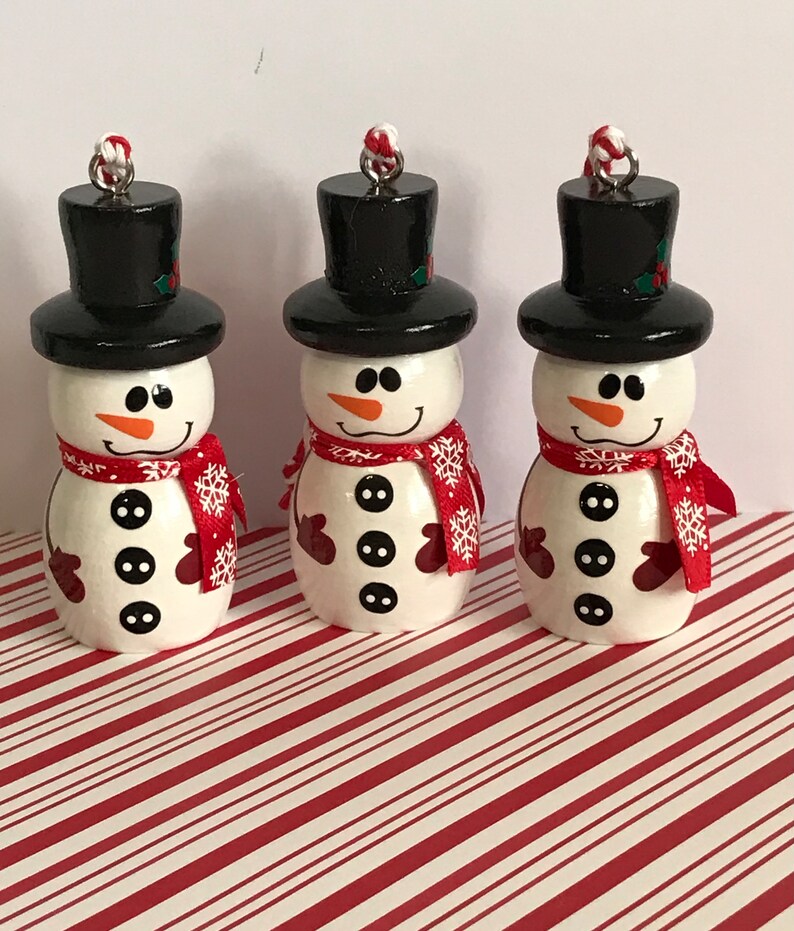 May include: Three small wooden snowman ornaments with black top hats, red and white scarves, and black buttons. The snowmen are on a red and white striped background.