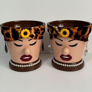May include: Two decorative terracotta flower pots painted with a woman's face. The pots have a brown rim and base, with a leopard print headband, a yellow sunflower, and pearl necklaces. Each pot has dangling earrings.