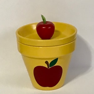 May include: A yellow ceramic planter with a red apple design. The planter has a separate top that can be used as a saucer. A small red apple sits on top of the saucer.