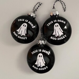 May include: Three black Christmas ornaments with the text "THIS IS SOME BOO SHEET" and a white ghost graphic. Each ornament has a silver cap and a black and white string for hanging. The ornaments are round and glossy.