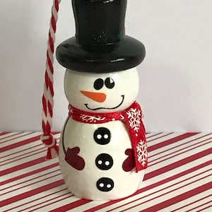 May include: A white snowman Christmas ornament with a black top hat, red scarf with white snowflakes, and black buttons. The snowman has a carrot nose and black eyes.