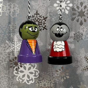 May include: Two Halloween-themed ornaments, one depicting Frankenstein's monster with green skin and a purple suit, and the other depicting a vampire with gray skin and a red suit.