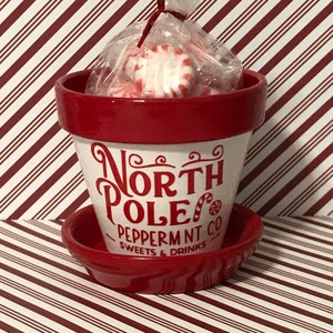 May include: A red and white ceramic planter with the words "North Pole Peppermint Co. Sweets & Drinks" written on it. The planter is filled with red and white peppermint candies.