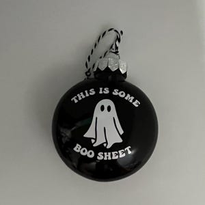 May include: Black round ornament with the text "THIS IS SOME BOO SHEET" and a white ghost illustration. The ornament has a silver cap and a black and white string for hanging. A Halloween-themed decoration.