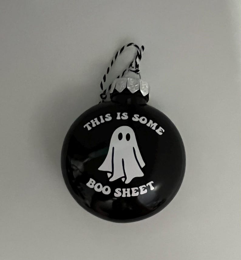 May include: Black round ornament with the text "THIS IS SOME BOO SHEET" and a white ghost illustration. The ornament has a silver cap and a black and white string for hanging. A Halloween-themed decoration.