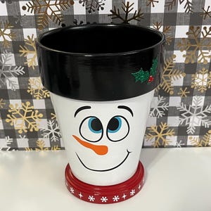 May include: A snowman-themed flower pot. The pot is white with a black rim and a red base. The snowman face has blue eyes, an orange carrot nose, and a smiling mouth. A sprig of holly is on the rim.