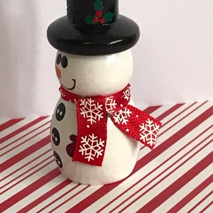 May include: A white snowman ornament with a black top hat, red and white snowflake patterned scarf, and black button eyes. The snowman is standing on a red and white striped surface.