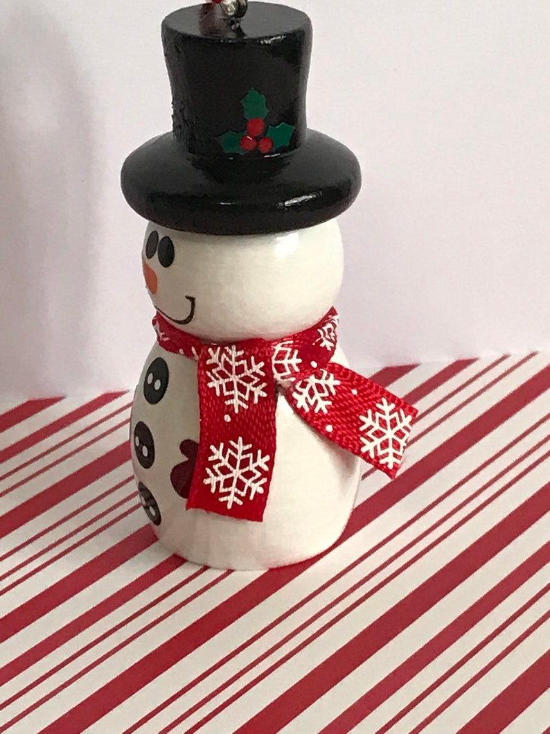 May include: A white snowman ornament with a black top hat, red and white snowflake patterned scarf, and black button eyes. The snowman is standing on a red and white striped surface.