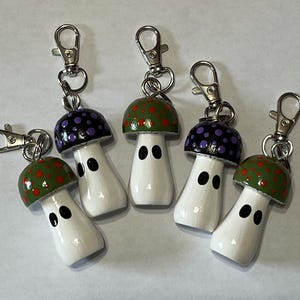 May include: Five mushroom-shaped keychains. Each keychain features a white body with two black oval eyes and a colorful cap. The caps are painted with either green and red spots or purple spots. Silver-colored clasps are attached.