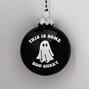 May include: Black round ornament with the text "THIS IS SOME BOO SHEET" in white. A white ghost graphic is in the center. The ornament has a silver metal cap and a black and white string for hanging.