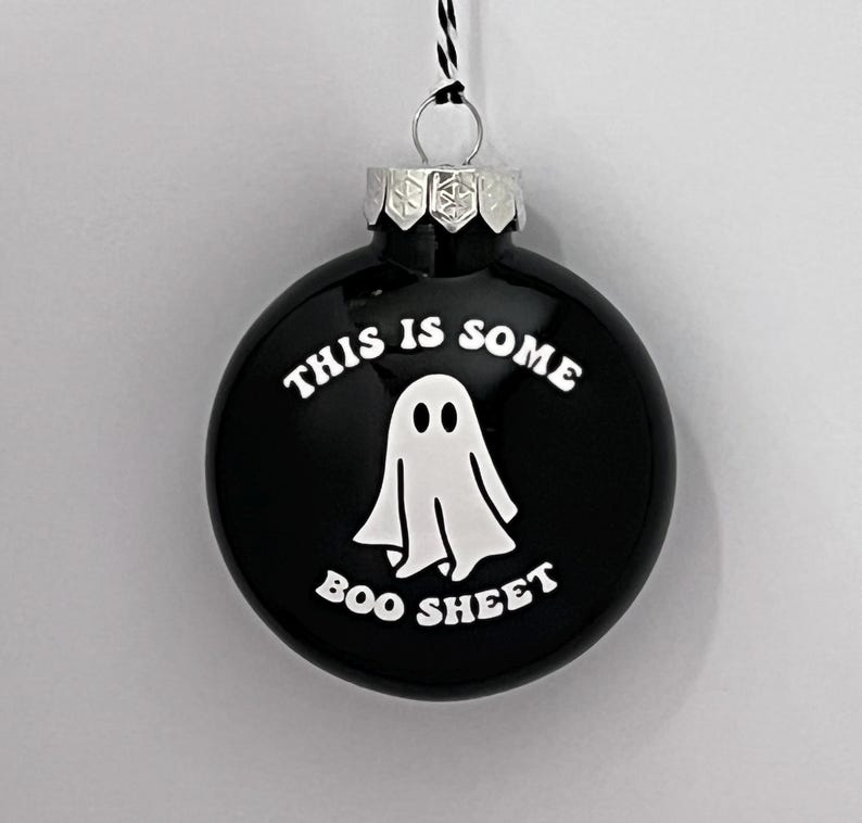 May include: Black round ornament with the text "THIS IS SOME BOO SHEET" in white. A white ghost graphic is in the center. The ornament has a silver metal cap and a black and white string for hanging.