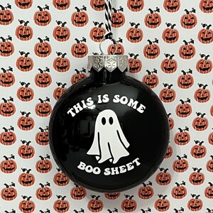May include: Black Christmas ornament with the text "THIS IS SOME BOO SHEET" and a white ghost graphic. The background features a pattern of orange jack-o'-lanterns on a white background. The ornament has a silver metal cap and a black and white string.