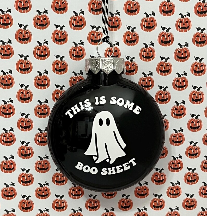 May include: Black Christmas ornament with the text "THIS IS SOME BOO SHEET" and a white ghost graphic. The background features a pattern of orange jack-o'-lanterns on a white background. The ornament has a silver metal cap and a black and white string.