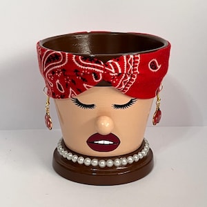May include: A decorative terracotta flower pot painted to resemble a woman's face. The pot features a red bandana, long eyelashes, a pearl necklace, and dangling earrings. The pot is brown inside and on the base.