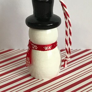 May include: A white snowman ornament with a black top hat and a red scarf with white snowflakes. The ornament has a white string for hanging.