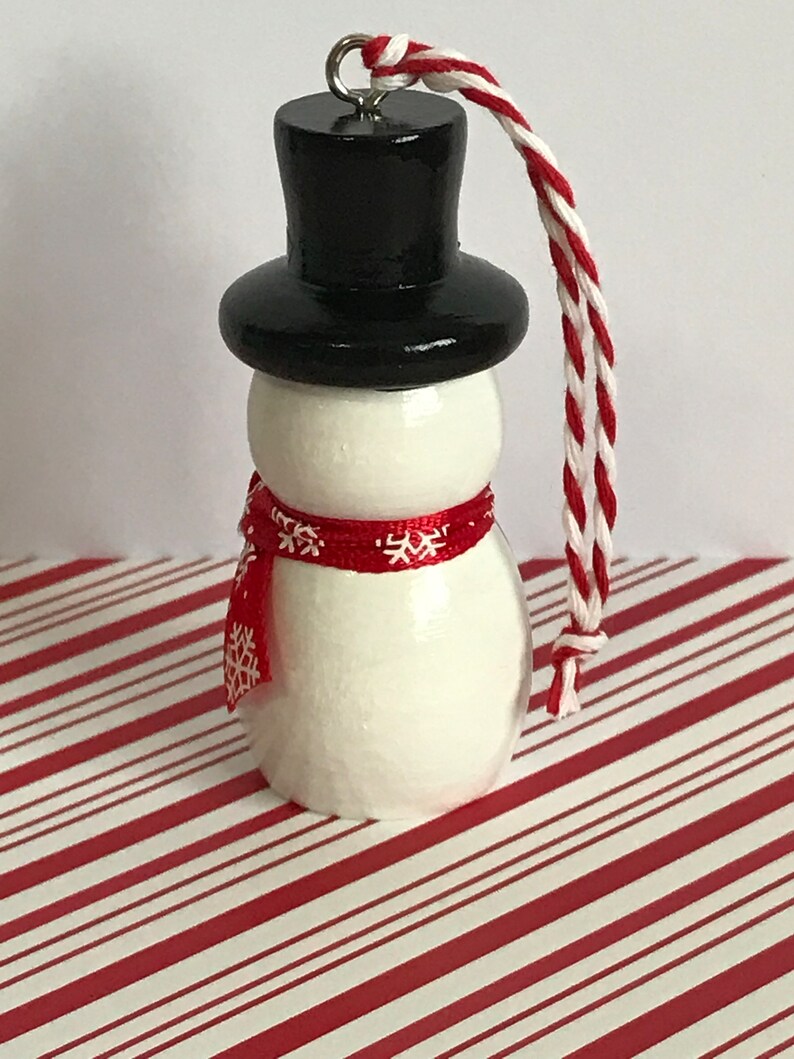 May include: A white snowman ornament with a black top hat and a red scarf with white snowflakes. The ornament has a white string for hanging.