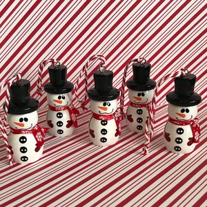 May include: Five white snowman ornaments with black hats, red scarves, and black buttons. The ornaments are hanging from twine and are against a red and white striped background.
