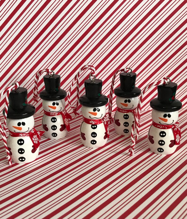 May include: Five white snowman ornaments with black hats, red scarves, and black buttons. The ornaments are hanging from twine and are against a red and white striped background.