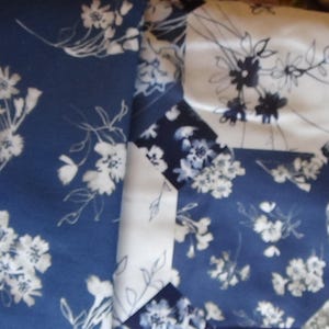 May include: Close-up of a patchwork textile featuring a floral pattern. The fabric combines navy blue, white, and light green sections, each adorned with delicate white floral designs. The overall aesthetic is reminiscent of a quilt or decorative fabric.