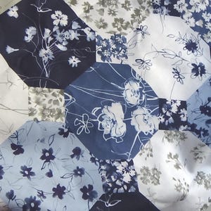 Può includere: Primo piano di una trapunta con motivo floreale. La trapunta presenta un design patchwork con forme esagonali in varie tonalità di blu, bianco e grigio. I motivi floreali includono fiori e foglie stilizzati.