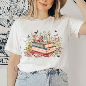 Puede incluir: Camiseta blanca con un gr&aacute;fico de una pila de libros rodeados de flores y mariposas. Los libros tienen un aspecto vintage y est&aacute;n decorados con una llave y un amuleto.