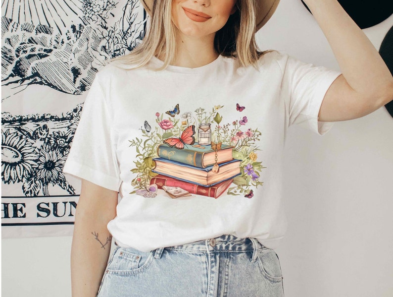 Puede incluir: Camiseta blanca con un gr&aacute;fico de una pila de libros rodeados de flores y mariposas. Los libros tienen un aspecto vintage y est&aacute;n decorados con una llave y un amuleto.