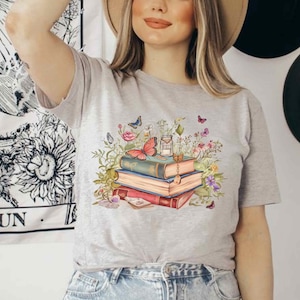 Puede incluir: Una camiseta gris con un gr&aacute;fico de una pila de libros, una mariposa y flores. Los libros est&aacute;n decorados con una llave y una botella de poci&oacute;n.