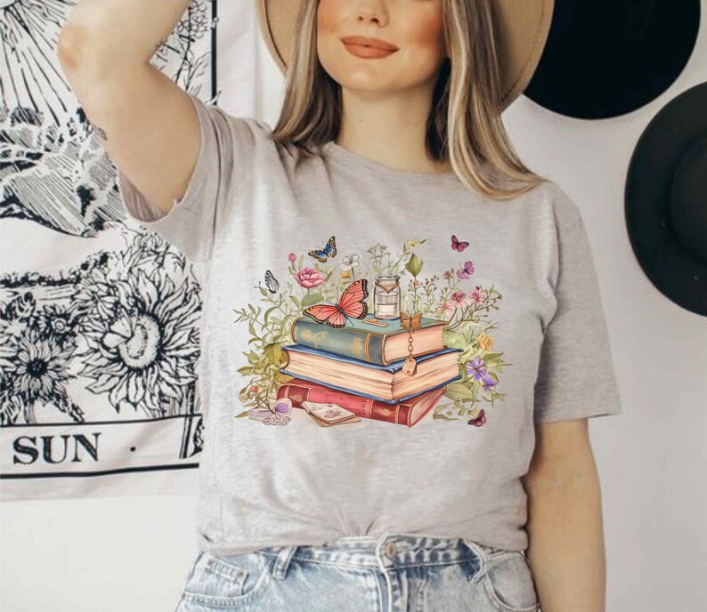 Puede incluir: Una camiseta gris con un gr&aacute;fico de una pila de libros, una mariposa y flores. Los libros est&aacute;n decorados con una llave y una botella de poci&oacute;n.