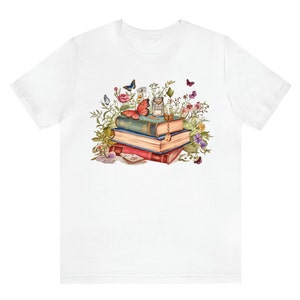 Puede incluir: Una camiseta blanca con una ilustraci&oacute;n colorida de una pila de libros vintage rodeados de flores y mariposas. Los libros est&aacute;n decorados con detalles dorados y una llave.