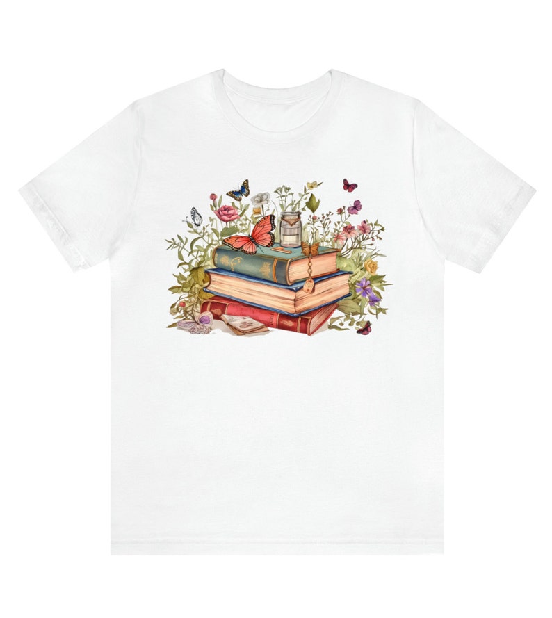 Puede incluir: Una camiseta blanca con una ilustraci&oacute;n colorida de una pila de libros vintage rodeados de flores y mariposas. Los libros est&aacute;n decorados con detalles dorados y una llave.