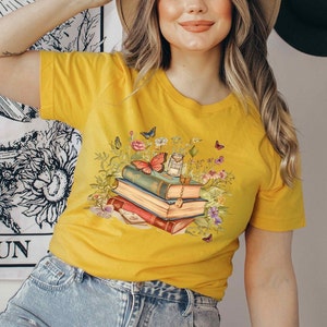 Puede incluir: Una camiseta amarilla con un gr&aacute;fico de una pila de libros con mariposas y flores. Los libros est&aacute;n apilados uno encima del otro y tienen diferentes colores y dise&ntilde;os.
