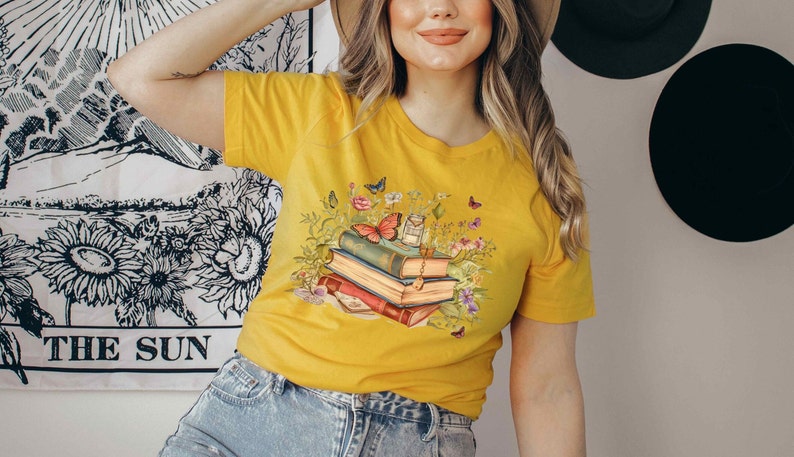 Puede incluir: Una camiseta amarilla con un gr&aacute;fico de una pila de libros con mariposas y flores. Los libros est&aacute;n apilados uno encima del otro y tienen diferentes colores y dise&ntilde;os.