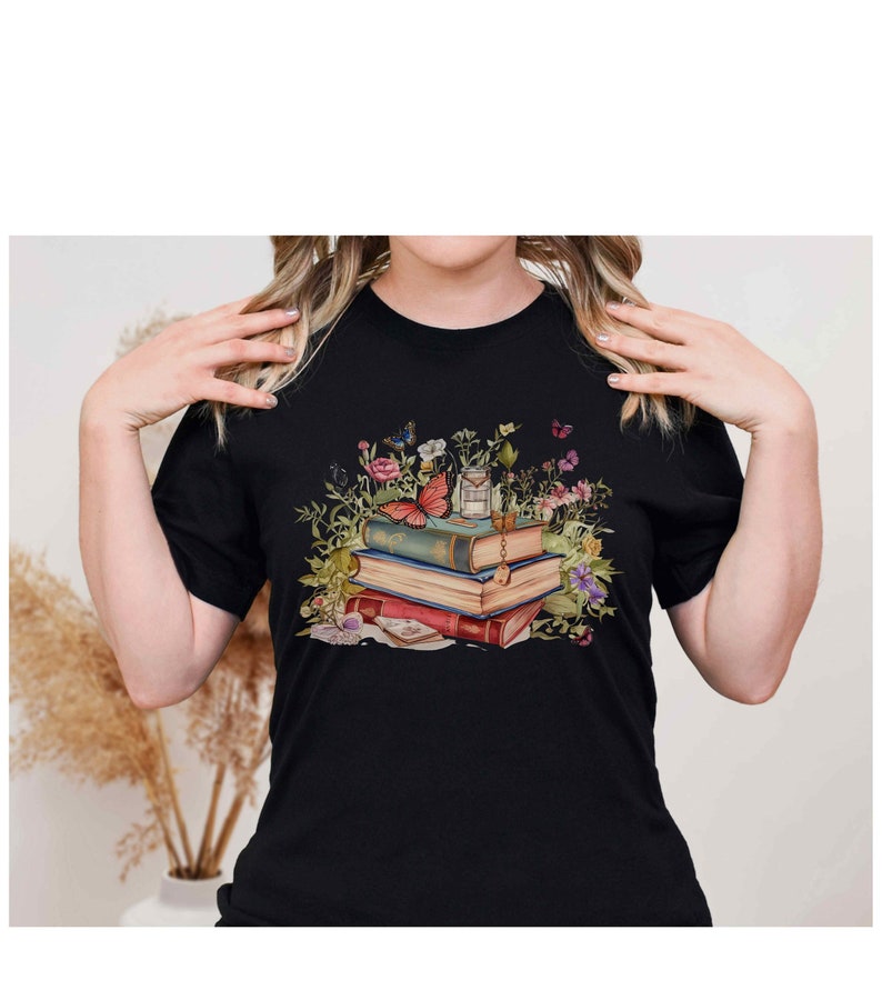 Puede incluir: Una camiseta negra con un gr&aacute;fico de una pila de libros antiguos rodeados de flores y mariposas. Un peque&ntilde;o frasco de vidrio con tapa de corcho se encuentra encima de los libros.