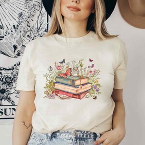 Puede incluir: Una camiseta blanca con un gr&aacute;fico de una pila de libros, flores y mariposas. Los libros est&aacute;n decorados con detalles dorados y las flores son de color rosa, morado y azul.