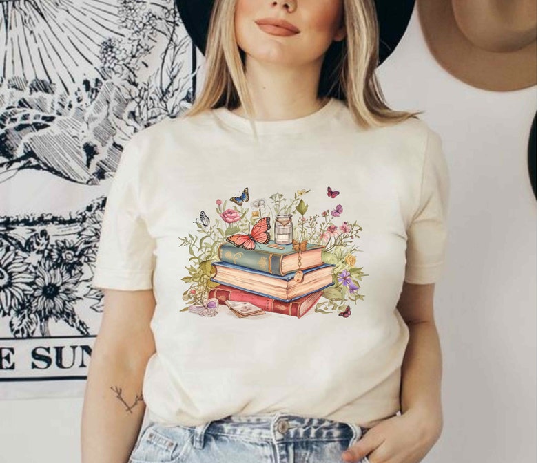 Puede incluir: Una camiseta blanca con un gr&aacute;fico de una pila de libros, flores y mariposas. Los libros est&aacute;n decorados con detalles dorados y las flores son de color rosa, morado y azul.