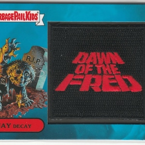 May include: A Garbage Pail Kids trading card featuring a rectangular black patch with red embroidered text reading DAWN OF THE FRED. To the left, an illustration of a zombie character named JAY DECAY stands by a tombstone marked R.I.P.
