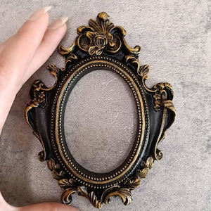 Può includere: Cornice ovale ornata, finitura nera e oro. La cornice presenta intricati dettagli floreali e a volute sui bordi. La cornice è tenuta in mano su uno sfondo neutro.