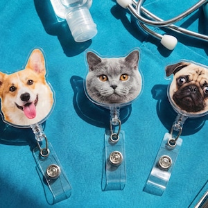 Puede incluir: Tres portatarjetas retr&aacute;ctiles con tem&aacute;tica de mascotas. Los dise&ntilde;os presentan un perro Corgi, un gato gris y un perro pug. Cada portatarjetas tiene una correa de pl&aacute;stico transparente y un broche met&aacute;lico. El fondo es una superficie azul.