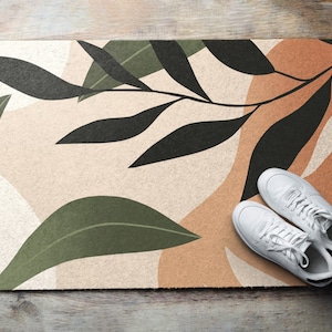 Peut inclure: Un paillasson beige avec un motif abstrait composé de feuilles noires et vertes. Le paillasson est de couleur marron, beige et blanc.
