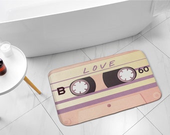 Könnte beinhalten: Eine rosa und gelbe Badematte mit einem Retro-Kassettenband-Design. Das Band hat die Worte "LOVE" und "60" aufgedruckt.