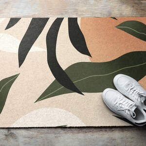 Peut inclure: Un paillasson beige avec un motif abstrait moderne composé de feuilles vertes et noires. Le paillasson est posé sur un sol en bois avec une paire de baskets blanches dessus.