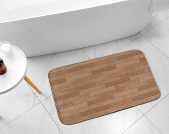 Könnte beinhalten: Eine rechteckige Badematte mit Holzmaserung in Brauntönen. Die Matte liegt auf einem weiß gefliesten Boden in der Nähe einer weißen Badewanne und eines kleinen Beistelltisches mit einer Kerze und einem Seifenspender.