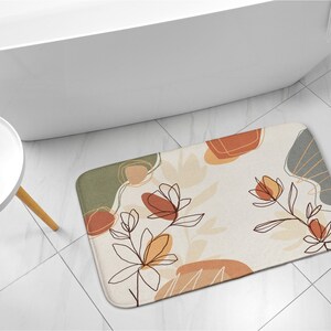 Op de afbeelding: Een beige badmat met een bloemenpatroon in tinten bruin, oranje en groen. Het patroon bevat abstracte vormen en lijnen.