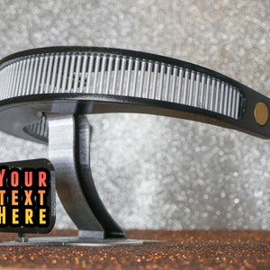 May include: A black and silver 3D printed object with a curved design. The object has a small sign attached to it that says "YOUR TEXT HERE" in red, orange, and yellow text.