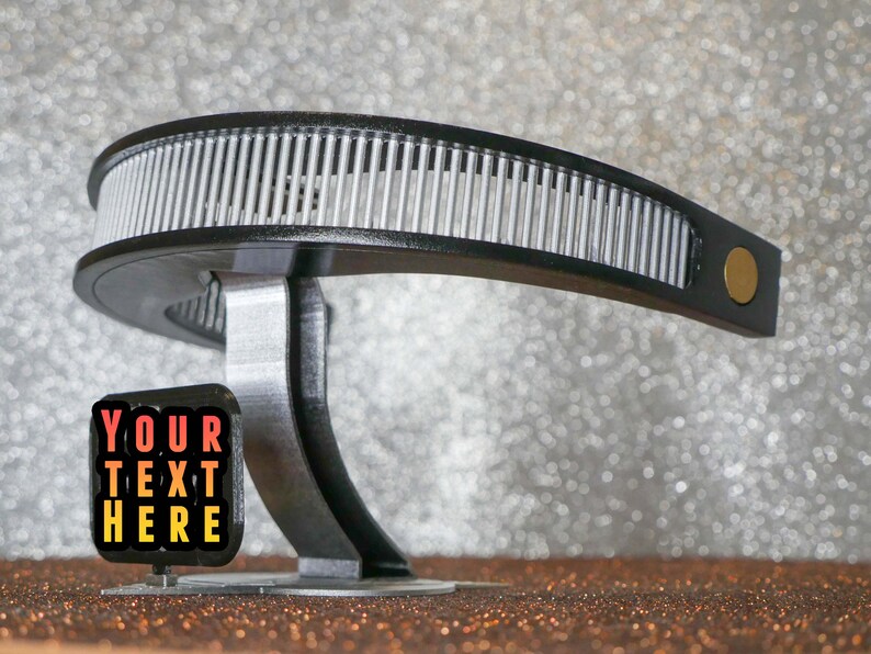 May include: A black and silver 3D printed object with a curved design. The object has a small sign attached to it that says "YOUR TEXT HERE" in red, orange, and yellow text.