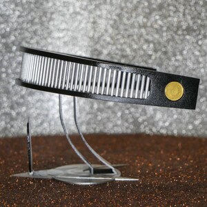 May include: A black and silver headband with a gold accent. The headband is on a stand with three legs.