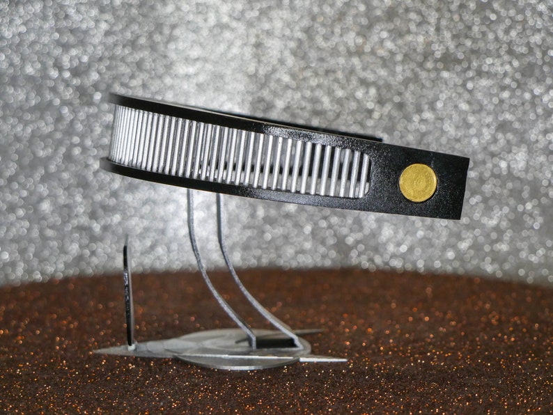 May include: A black and silver headband with a gold accent. The headband is on a stand with three legs.