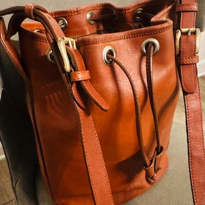 Puede incluir: Un bolso tipo cubo de cuero marrón con cierre de cordón y correa ajustable para el hombro. El bolso presenta herrajes de color latón, incluyendo hebillas y ojales. El cuero tiene un tono cálido y rico.